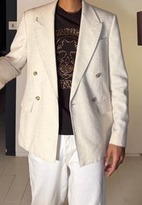 Personne portant un blazer beige croisé sur une chemise noire ornée d'un crâne clouté doré et un pantalon blanc, debout à l'intérieur.
