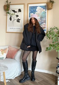 Nafouknutá černá vesta přes tmavě šedou mikinu a minisukni, doplněná černými vysokými botami. Měkká růžová čepice a kudrnaté vlasy dokončují vzhled.