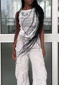 Γκρίζο γραφικό αμάνικο μπλουζάκι με στρογγυλή λαιμόκοψη; μπεζ παντελόνι cargo με πλαϊνές τσέπες; μακριές μαύρες πλεξούδες; φόντο σε φυσικές αποχρώσεις.