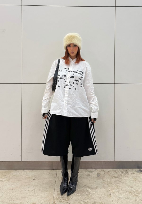 Asmuo dėvi baltą marškinėlį su spaudiniu, juodus plačius Adidas šortus, juodus smailius batus, kreminę pūkuotą kepurę ir neša juodą petinę rankinę.