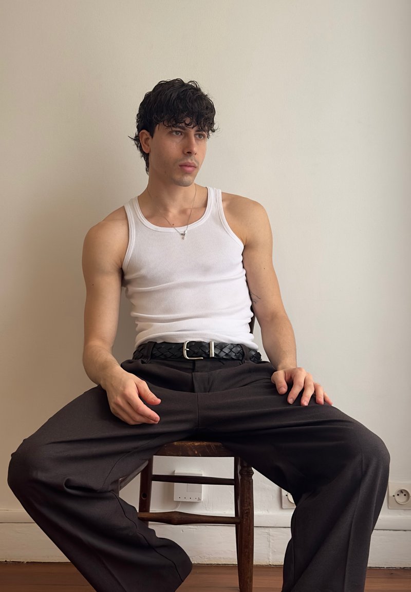 Young man with curly dark hair wearing a white tank top and black pants, sitting on a wooden chair against a plain beige wall.