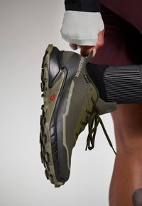 Olivovo zelená bežecká obuv na trail s textúrovaným sieťovinovým zvrškom, čiernou gumovou podrážkou, červeným akcentom a zakriveným dizajnom päty. Systém šnurovania.