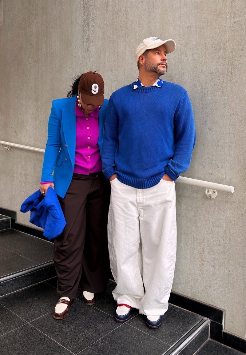 Deux personnes se tiennent sur des escaliers près d'un mur en béton ; l'une porte un pull bleu et un pantalon blanc, l'autre un blazer bleu, une chemise rose et une casquette marron.