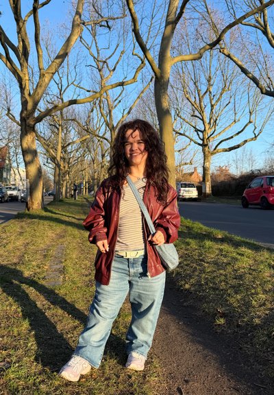 Joven con cabello rizado, lleva una chaqueta roja y jeans, está de pie sonriendo en un camino bordeado de árboles junto a una carretera, bajo la luz del atardecer.