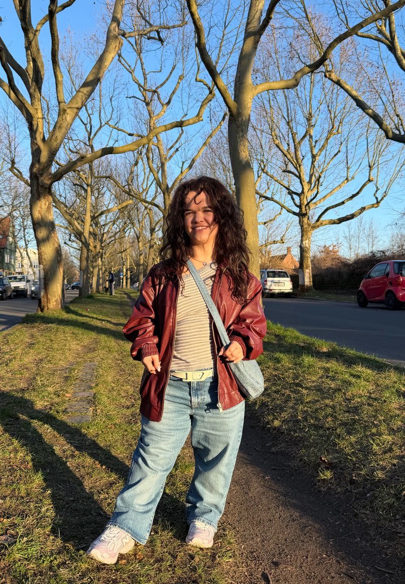 Jeune femme aux cheveux bouclés portant une veste rouge et un jean, souriante, debout sur un chemin bordé d'arbres à côté d'une route, à la lumière de la fin d'après-midi.