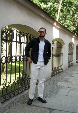 Chaqueta negra de botones, camiseta blanca y pantalones crema. Los accesorios incluyen gafas de sol naranjas y zapatos negros. Escenario al aire libre con un camino de piedra.