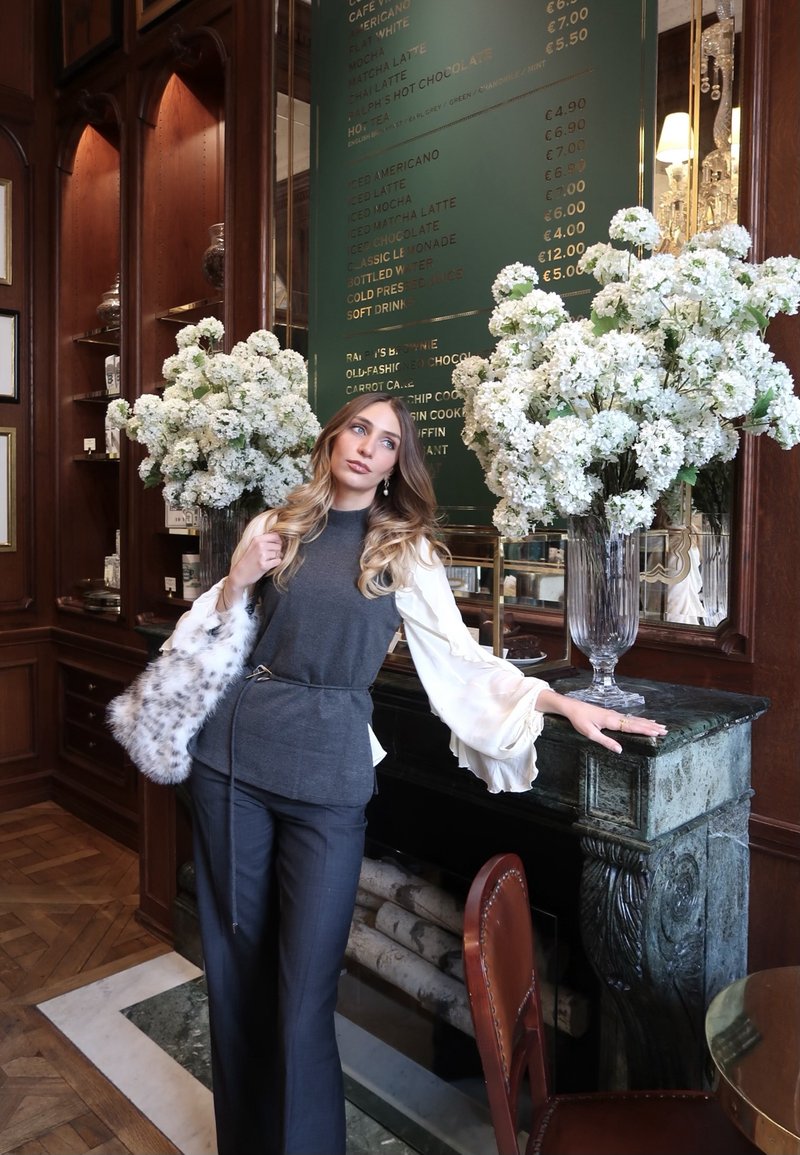 Γυναίκα με μακριά μαλλιά και λευκό πουκάμισο γέρνει σε ένα πράσινο μαρμάρινο τζάκι, δίπλα σε μεγάλους βάζους με λευκά λουλούδια σε ένα δωμάτιο με ξύλινες επενδύσεις.