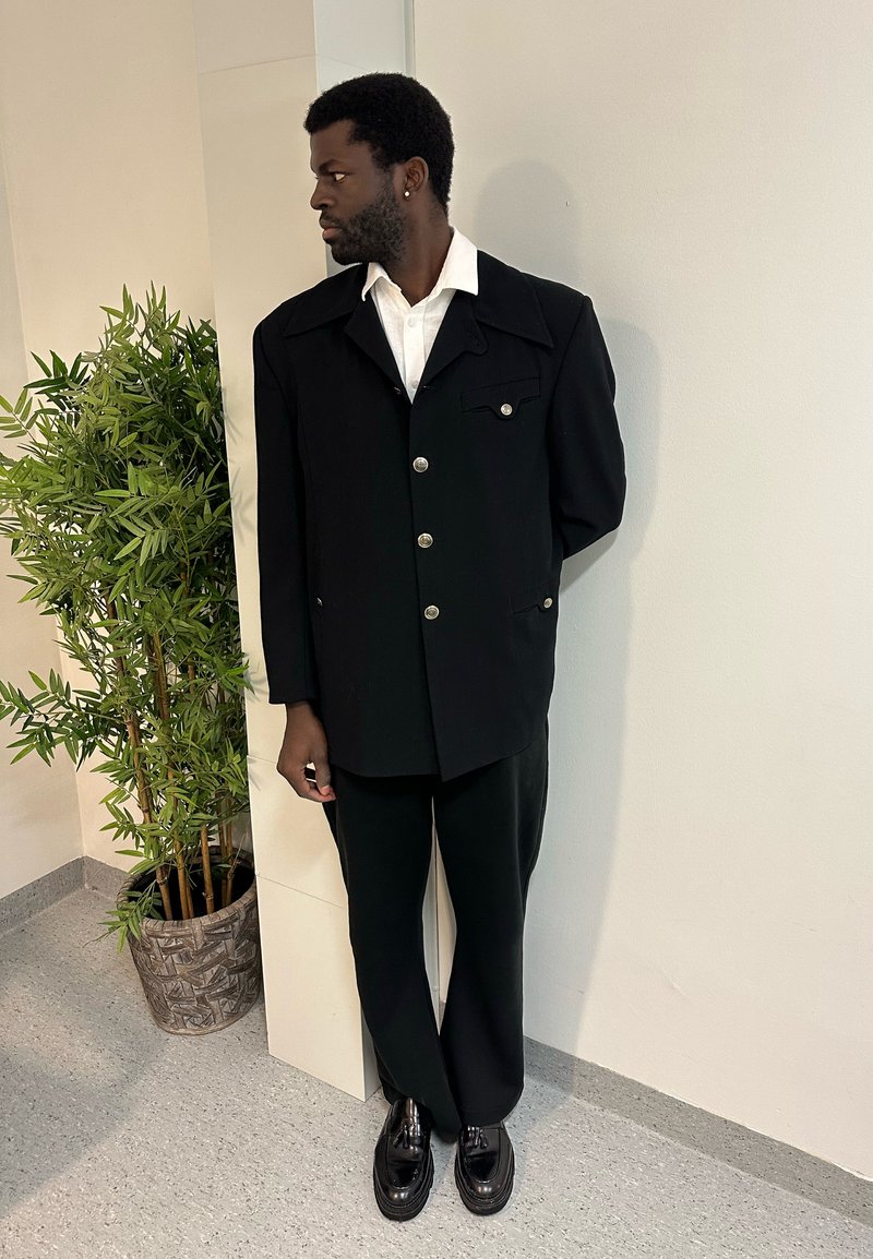 Άνδρας με μαύρο κοστούμι και άσπρο πουκάμισο, στέκεται σε εσωτερικό χώρο κοντά σε ψηλό φυτευμένο φυτό, κοιτώντας προς τα αριστερά με τα χέρια πίσω από την πλάτη.