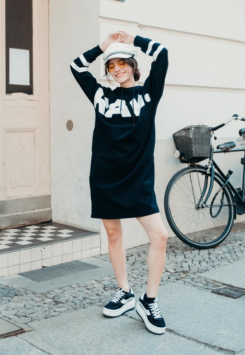 Fekete kapucnis ruha fehér grafikai szöveggel, hosszú ujjakkal, fehér csíkokkal, fekete-fehér sportcipővel párosítva. Városi háttér előtt.