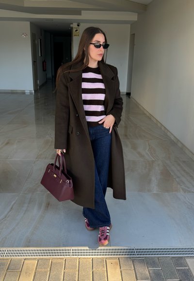 Mujer con abrigo marrón oscuro, suéter a rayas, jeans anchos azules, zapatillas rosas y gafas de sol, sosteniendo un bolso burdeos en interiores.