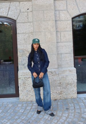 Dunkelblaue Jeanshemd mit Knöpfen, weit geschnittene blaue Jeans, schwarze geflochtene Handtasche, grüne Mütze mit weißem Logo, schwarze Slippers mit weißen Punkten.