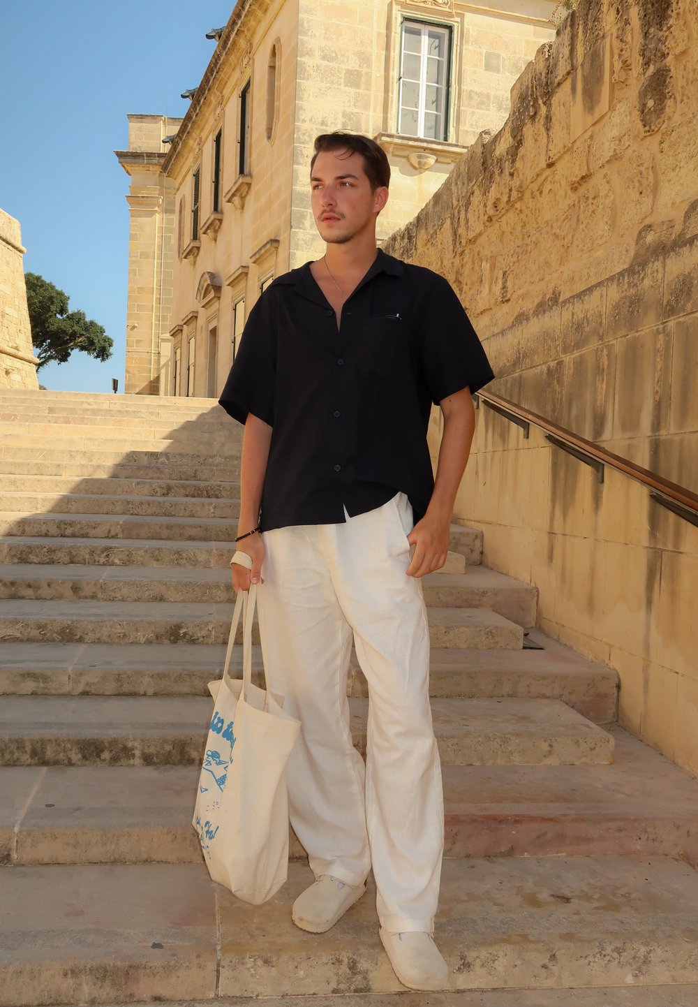 Zwarte short-sleeve shirt, witte lichte broek, een beige schoudertas met blauw patroon dragend, staand op stenen trappen. Natuurlijk licht.