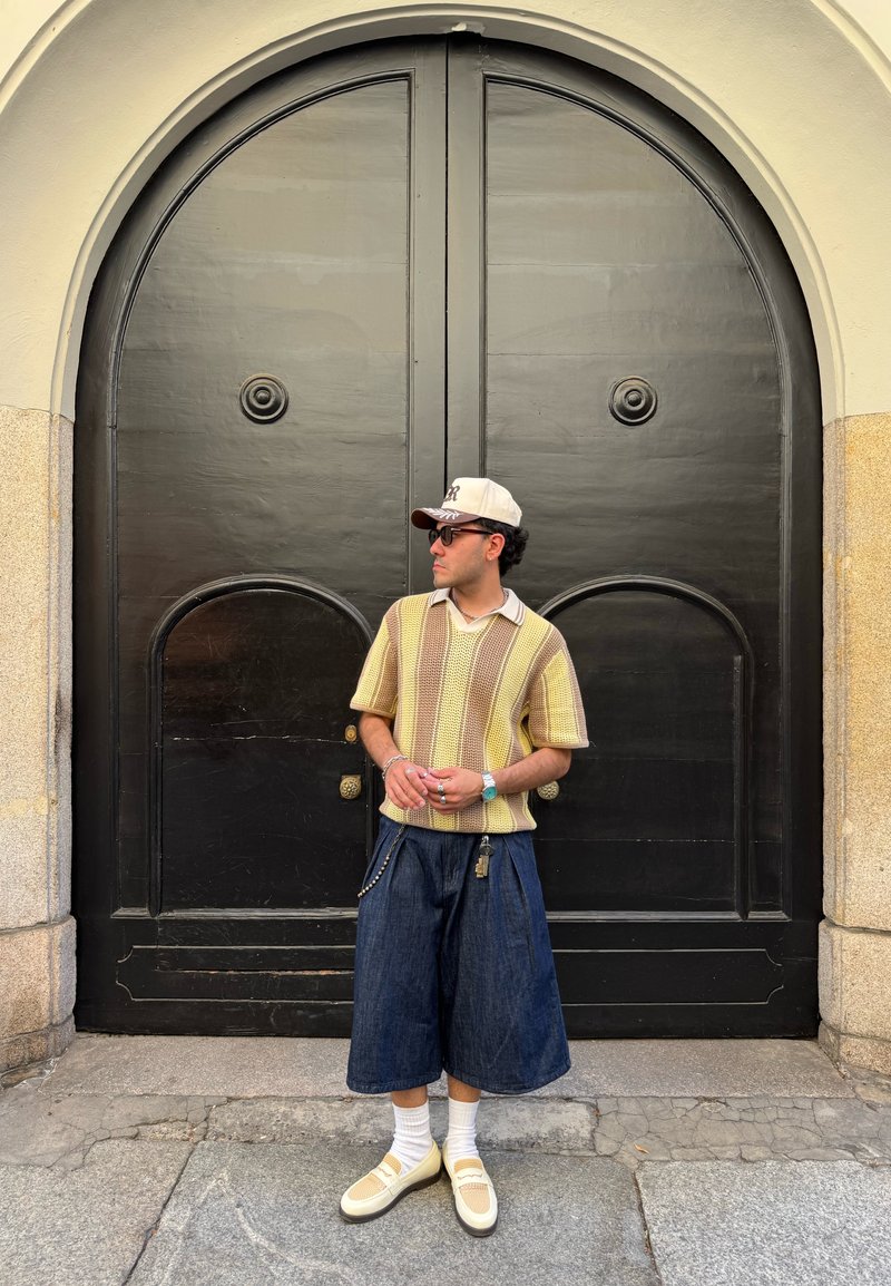 Chandail à manches courtes en rayures jaunes et brunes, culottes en denim foncé, chaussettes blanches, baskets jaunes, bijoux en argent et casquette beige.