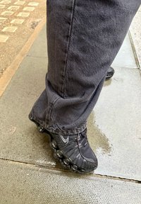 Personne portant des baskets Nike noires avec des semelles à bulles uniques et un pantalon foncé, debout sur un trottoir mouillé en plein air.