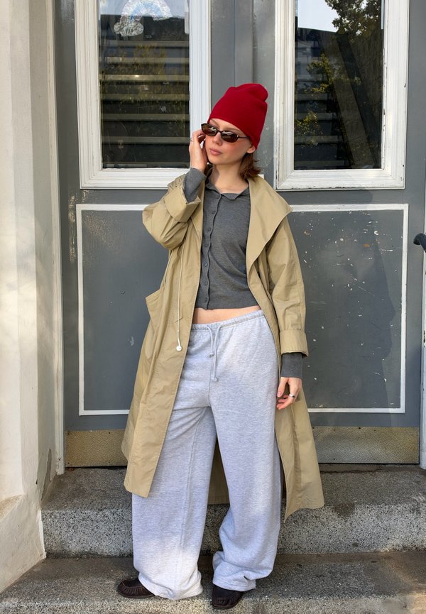 Une femme portant un bonnet rouge, des lunettes de soleil, un trench beige, un haut gris et un pantalon de jogging ample ajuste ses lunettes à l’extérieur, devant une fenêtre.