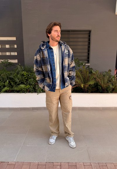 Hombre con las manos en los bolsillos, lleva una chaqueta de cuadros sobre una camisa de mezclilla y pantalones cargo beige, de pie sobre un suelo de baldosas cerca de plantas y una pared gris.