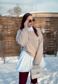 Femme en tenue d'hiver en extérieur tenant un sac à main beige, ajustant ses lunettes de soleil, avec une clôture en bois et un sol enneigé en arrière-plan.