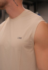 Camiseta atlética sem mangas bege, feita de tecido texturizado, com um pequeno logótipo "ICIW" no lado esquerdo da frente.