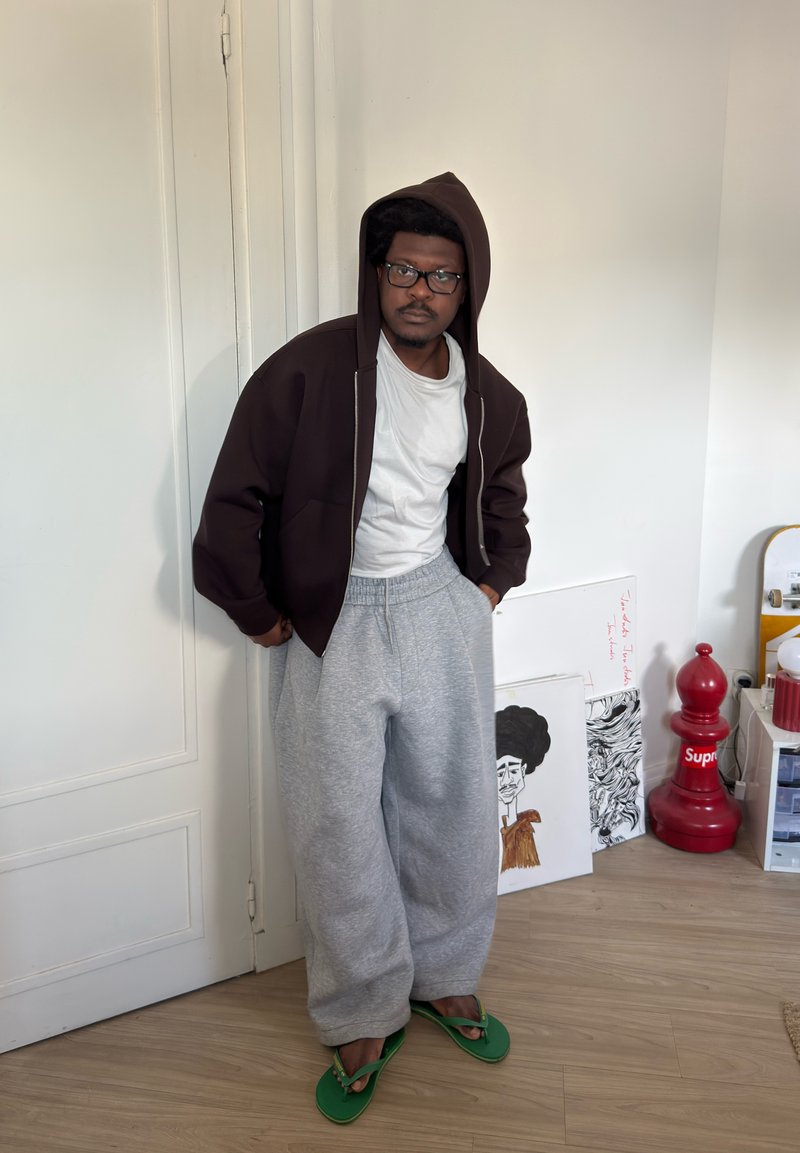 Man som bär glasögon, brun hoodie, vit t-shirt, gråa mjukisbyxor och gröna flip-flops står inomhus bredvid konstverk och en stor röd schackpjäs.