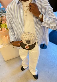 Chemise blanche superposée d'une surchemise à rayures bleues, pantalon blanc, chaussures noires, bijoux en or et casquette de baseball grise avec un logo.