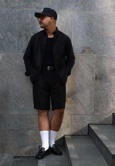 Camisa negra de manga corta sobre una camiseta negra, combinada con pantalones cortos negros y mocasines negros. Medias blancas hasta el tobillo completan el atuendo frente a un fondo de piedra.