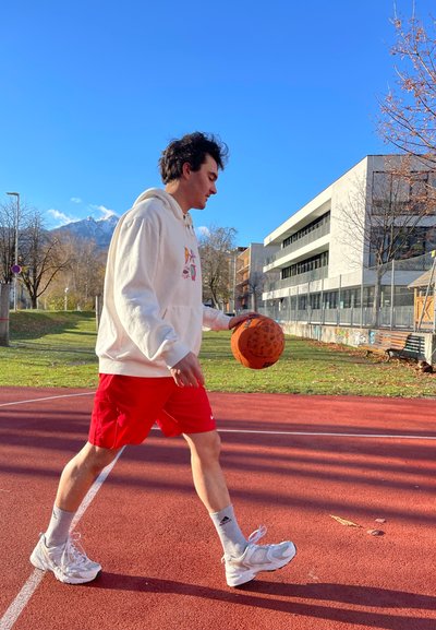 Una persona con una sudadera blanca y pantalones cortos rojos dribla un baloncesto en una cancha roja, con edificios y árboles en un fondo soleado.