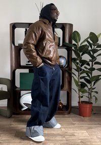 Blouson aviateur en cuir marron avec poignets côtelés, jean oversize bleu foncé, et chaussures avec accents gris. Intérieur avec des plantes en arrière-plan.