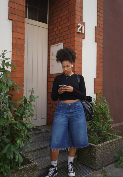 Top negro de manga larga, culottes de mezclilla azul oversized con dobladillo deshilachado, zapatillas negras, sosteniendo un teléfono inteligente, cerca de un edificio de ladrillo.