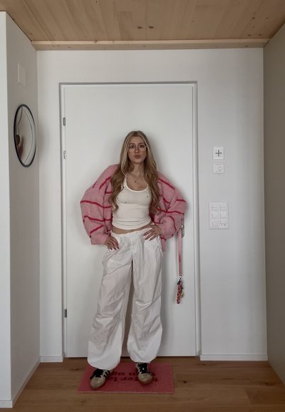 Joven mujer con cabello rubio largo, que lleva gafas, cárdigan de rayas rosas, camiseta sin mangas blanca, pantalones blancos y zapatillas, de pie dentro de una habitación junto a una puerta cerrada.