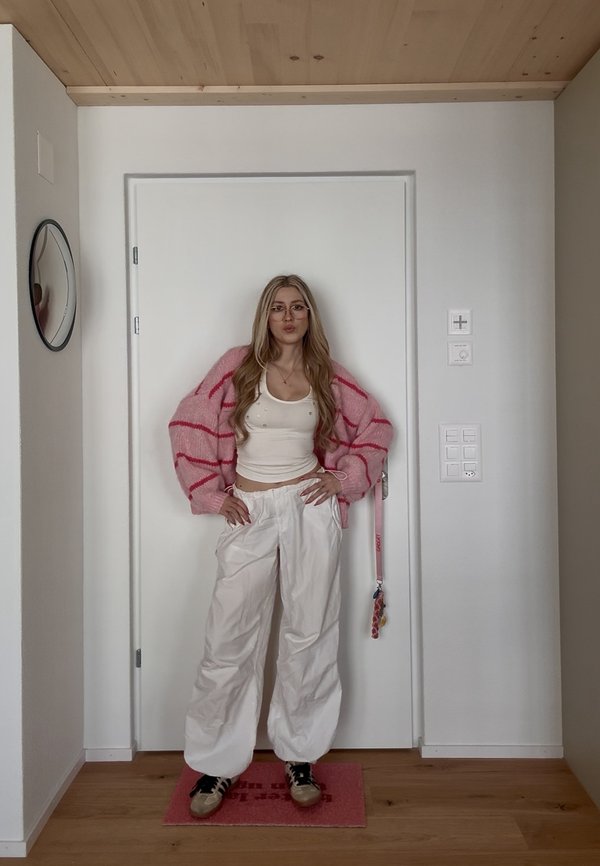 Mladá žena s dlouhými blond vlasy nosící brýle, růžový pruhovaný cardigan, bílý tílko, bílé kalhoty a tenisky stojí uvnitř u zavřených dveří.