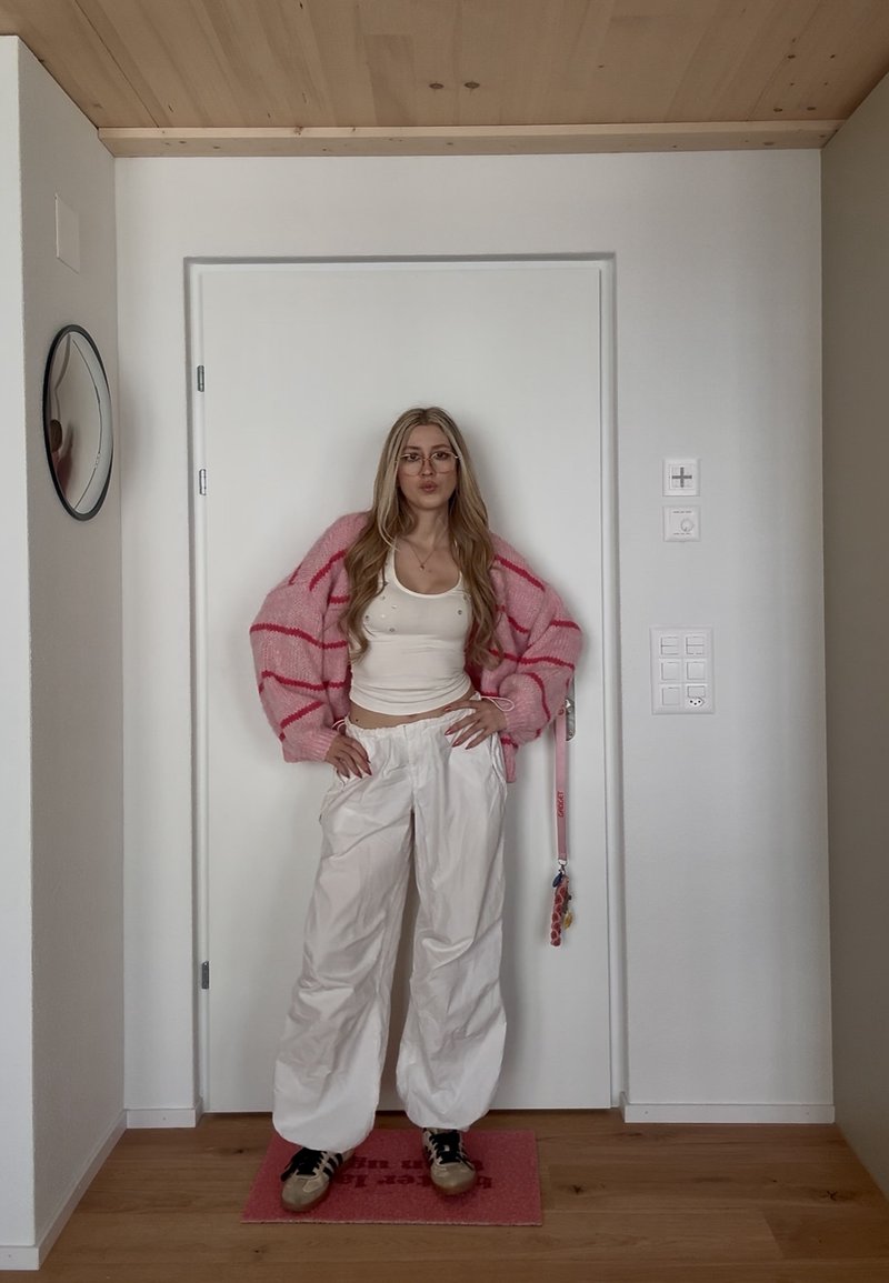 Junge Frau mit langen blonden Haaren, die eine Brille, einen rosa gestreiften Cardigan, ein weißes Tanktop, weiße Hosen und Turnschuhe trägt, steht drinnen an einer geschlossenen Tür.