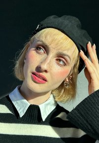 Μαύρο μπερέ, λευκό πουκάμισο με γιακά και μαύρο-άσπρη ριγέ πουλόβερ. Φακίδες, ελαφρύ μακιγιάζ και ασημένια σκουλαρίκια κρίκοι. Μαλακό, υφασμάτινο υλικό.