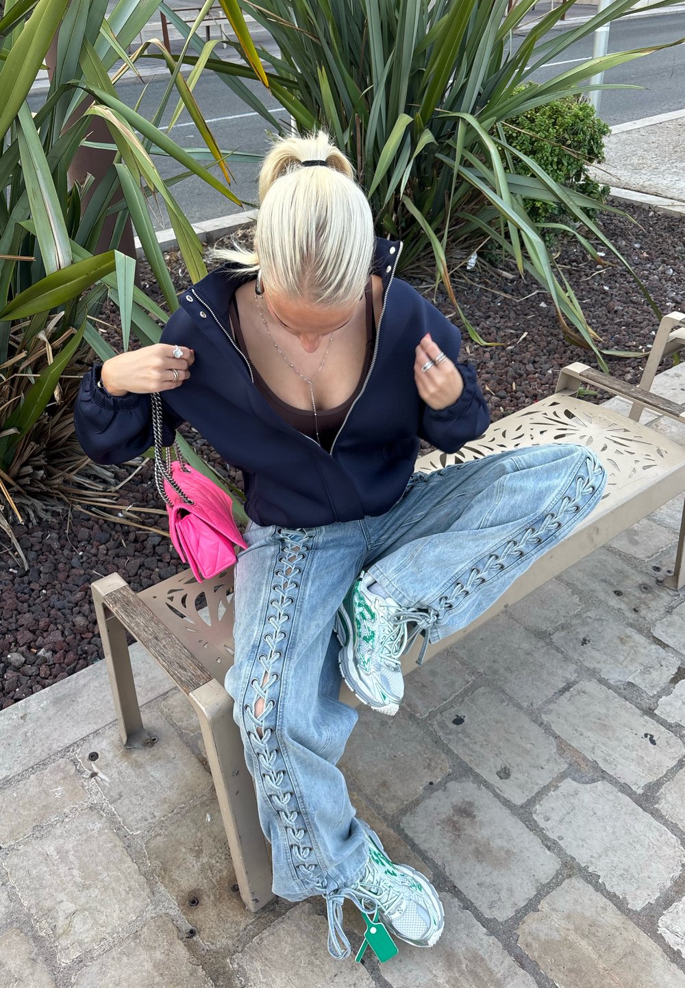 Plave osobe s repom sjedi prekriženih nogu na klupi, odjevene u tamnoplavu jaknu, traperice s vezicama, bijele tenisice i drži svijetlu ružičastu torbu.