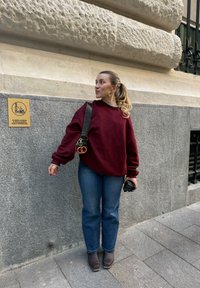 Sweatshirt oversized bordeaux, suéter leve por baixo, jeans azuis, botas castanhas até ao tornozelo e câmara preta. Placa na parede atrás.