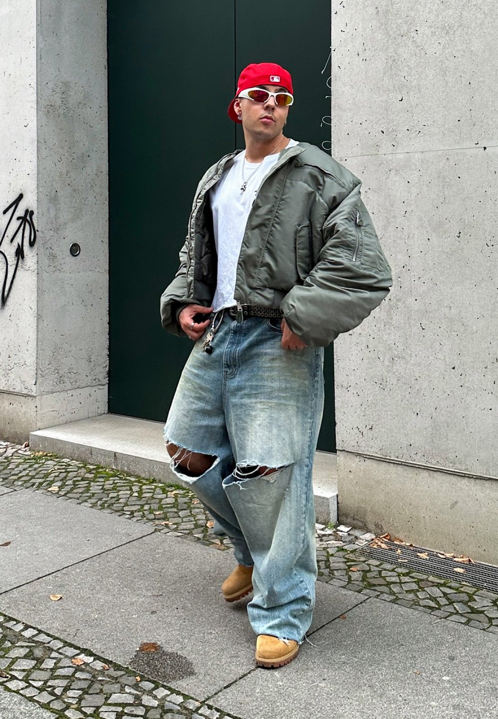 Chaqueta de pluma verde, camiseta blanca, jeans claros desgastados con grandes rotos, calzado beige, gorra roja y gafas de sol. Entorno urbano.