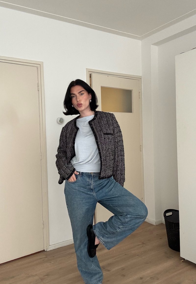 Femme aux cheveux noirs debout à l'intérieur, en équilibre sur une jambe, portant un haut gris, une veste à motif, un jean large et des chaussures noires.