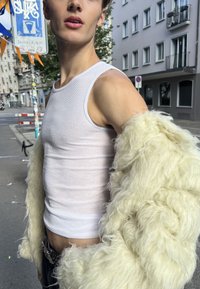 Jeune personne portant un débardeur blanc et une veste crème duveteuse surdimensionnée, debout dans une rue de la ville avec des bâtiments et des enseignes en arrière-plan.