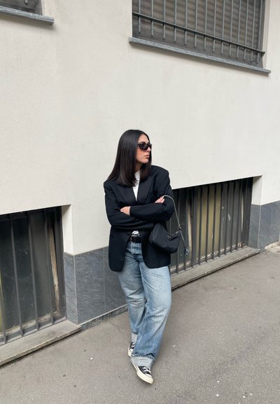 Blazer negro sobre una camisa blanca, jeans holgados de color azul claro y zapatillas negras. Lleva un pequeño bolso negro. Cabello largo y liso, usando gafas de sol.