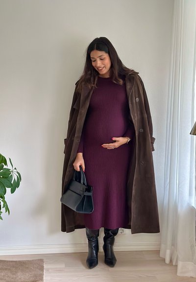 Vestido premamá de canalé en burdeos, abrigo largo de ante marrón, botas negras de tacón, sosteniendo un pequeño bolso negro, de pie contra una pared clara.