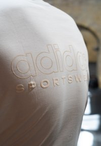 Lys beige bomulds T-shirt med præget "adidas SPORTSWEAR" logo. Blød tekstur med korte ærmer, minimalistisk design.