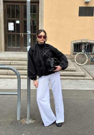 Mujer con chaqueta negra de gran tamaño, pantalones blancos de pierna ancha y gafas de sol, sosteniendo un bolso negro, de pie en la acera de la ciudad cerca de unas escaleras y una bicicleta.