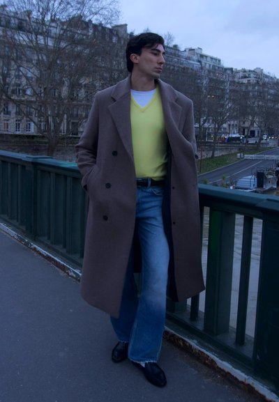 Joven con abrigo largo marrón, suéter amarillo, vaqueros azules y zapatos negros apoyado en la barandilla verde de un puente junto a un río al anochecer.