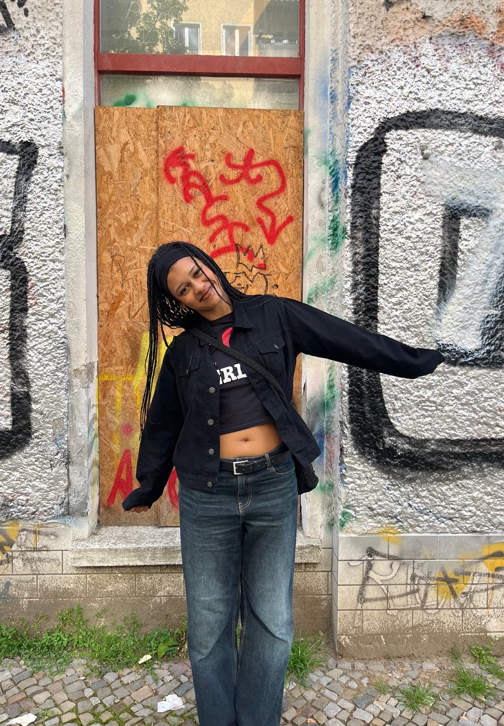 Black denim jacket over a black crop top and blue jeans. Long braids, standing against a distressed wall with graffiti and wooden boards.