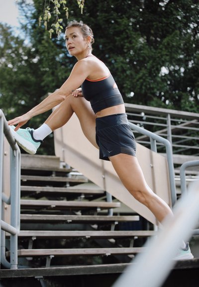 Atleta en un sujetador deportivo negro y pantalones cortos estirándose en las escaleras. Lleva zapatillas turquesas y calcetines blancos, con el cabello recogido.