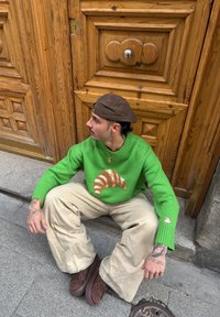 Νέος άνδρας με πράσινο πουλόβερ με σχέδιο κρουασάν, μπεζ παντελόνι και καφέ παπούτσια, καθισμένος σε πεζοδρόμιο μπροστά σε ξύλινη πόρτα.