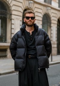 Doudoune noire avec un col montant et des détails zippés, associée à une chemise noire et un pantalon noir ample. Comprend des lunettes de soleil.