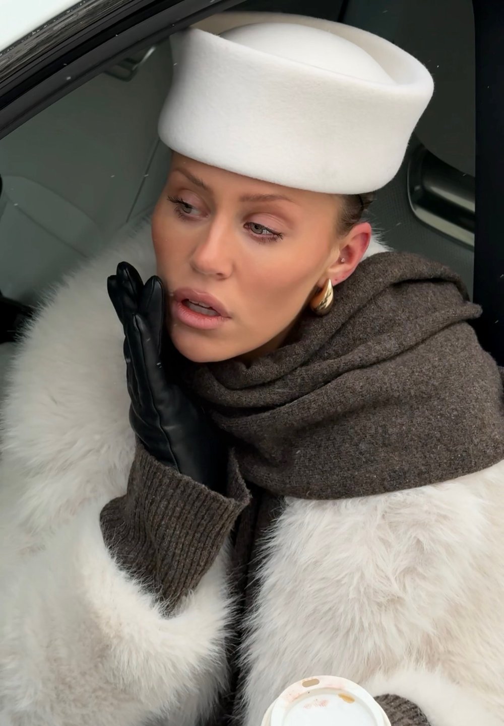 Femme portant un chapeau blanc, un manteau en fourrure, une écharpe marron et des gants noirs, tenant une tasse de café, appuyant son visage sur sa main gantée à l'intérieur d'une voiture.