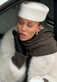 Femme portant un chapeau blanc, un manteau en fourrure, une écharpe marron et des gants noirs, tenant une tasse de café, appuyant son visage sur sa main gantée à l'intérieur d'une voiture.