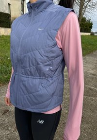 Lilla vattert vest med høy krage, glidelåslukking og elastisk midjebånd over en lys rosa langermet topp og svarte leggings med logoer.