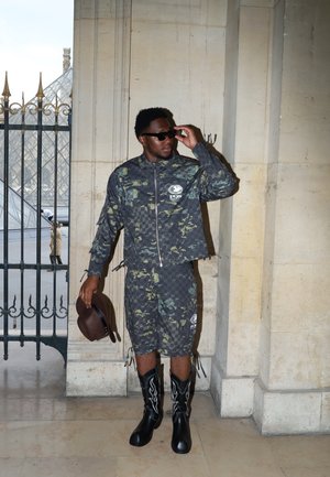 Chaqueta y pantalones cortos de camuflaje en tonos de verde y gris, llevados con botas vaqueras negras con acentos de llamas blancas. Sosteniendo un sombrero marrón.